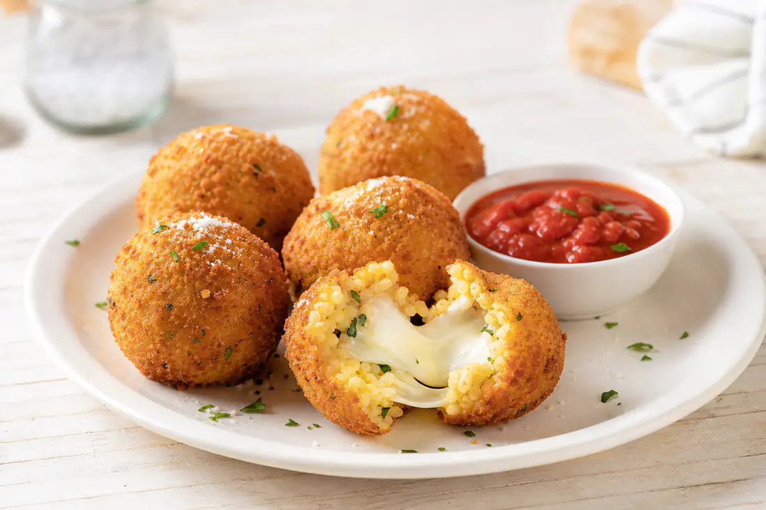 Round breaded arancini, one cut open to show melted mozzarella, with marinara sauce
