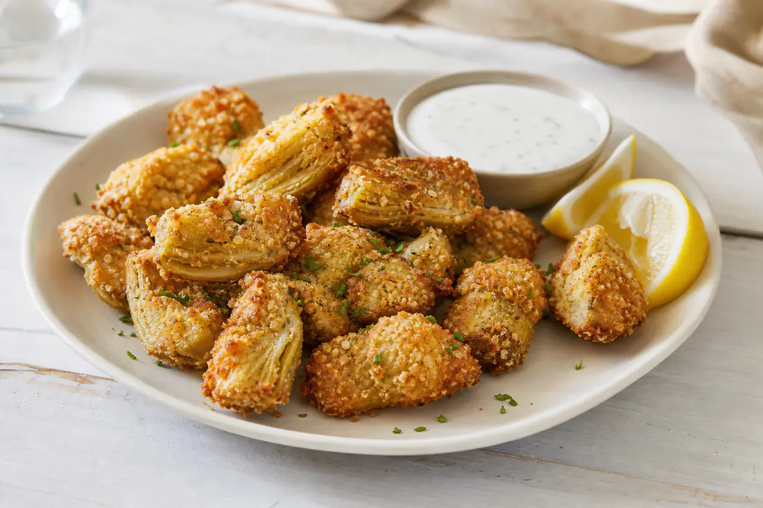 Golden breaded artichoke heart quarters on a plate with a bowl of ranch dip and lemon wedges
