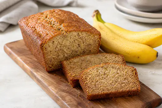 Sliced banana bread in a small LOAF pan, with a golden brown crust and moist crumb, next to two ripe bananas