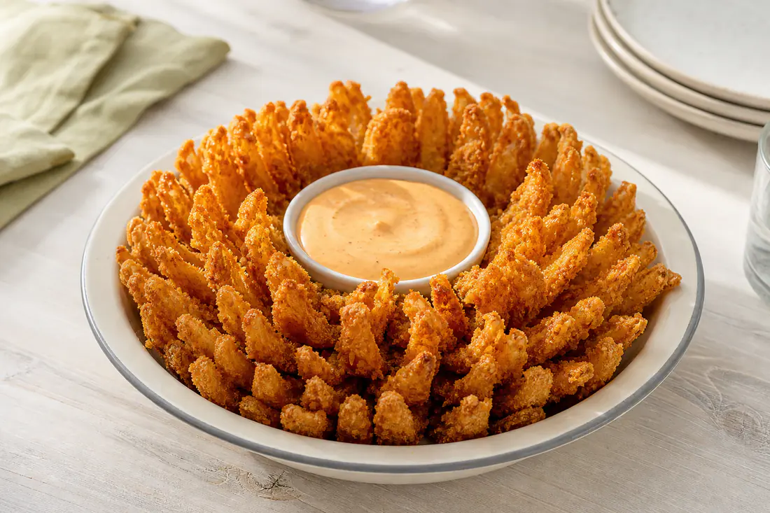 A golden panko-breaded blooming onion with open petals in a bowl, next to a creamy dipping sauce
