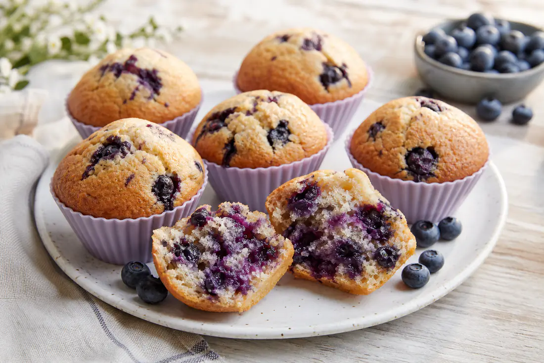 Golden blueberry muffins with domed tops in silicone cups, with fresh blueberries scattered beside them