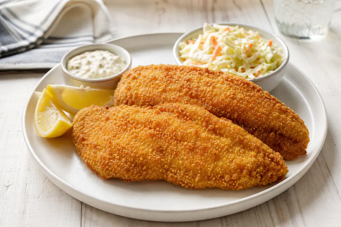 Golden cornmeal-crusted catfish fillets on a plate with lemon wedges and tartar sauce