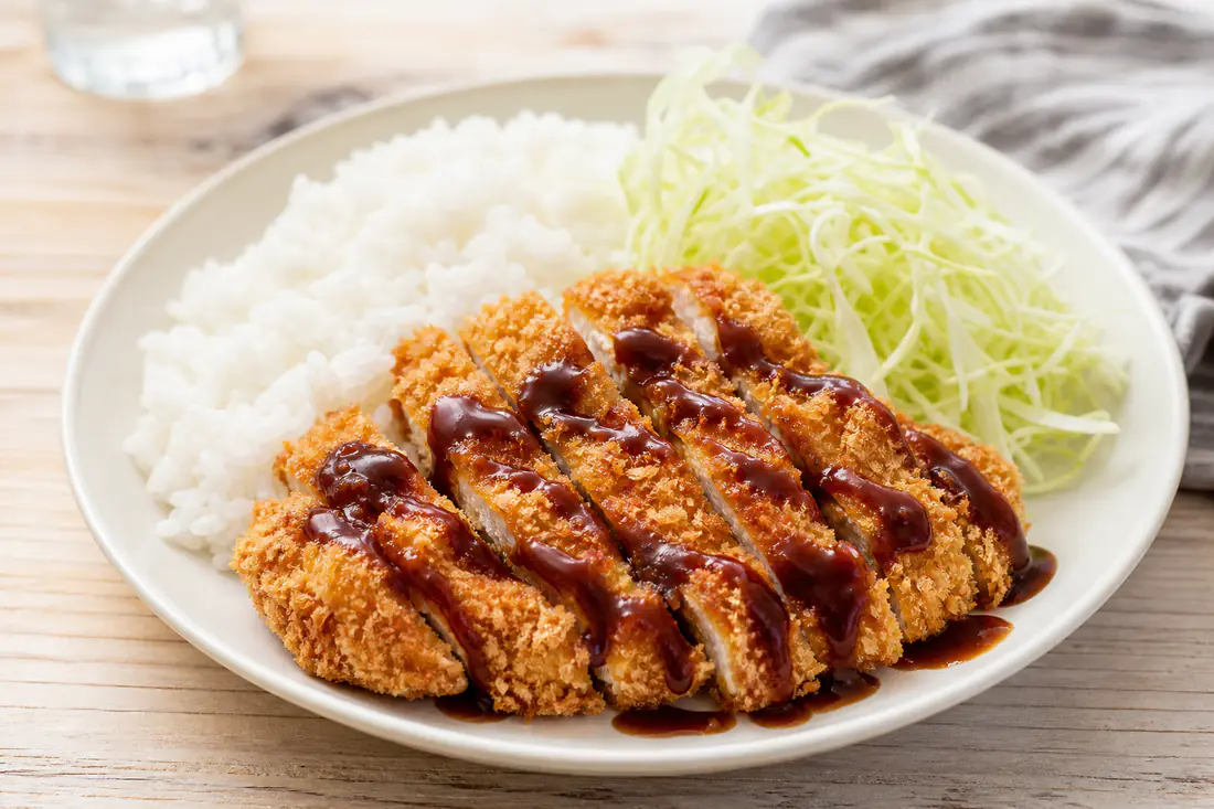 Sliced golden chicken katsu cutlets on a plate with white rice, shredded cabbage and katsu sauce