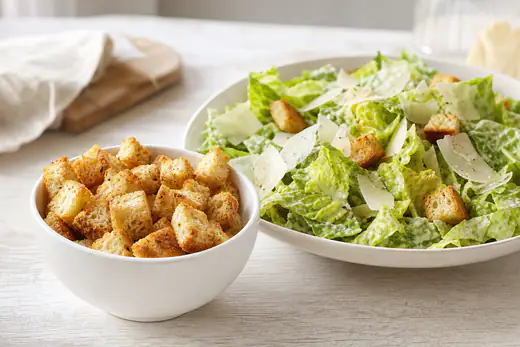 Golden homemade croutons in a white bowl next to a Caesar salad