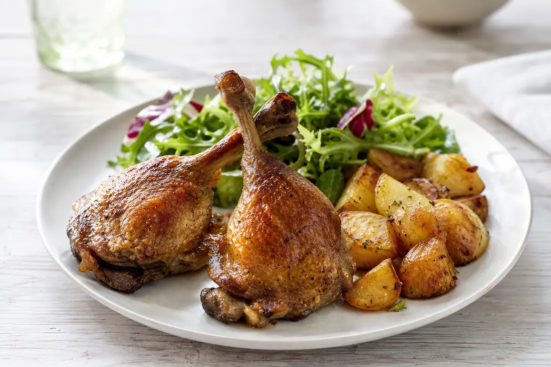 Two crispy roasted duck legs with golden brown skin on a plate with roasted potatoes