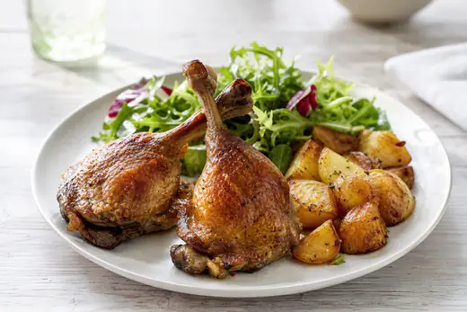 Two crispy roasted duck legs with golden brown skin on a plate with roasted potatoes