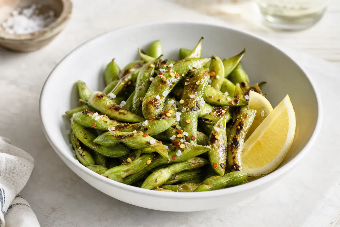 Blistered edamame pods sprinkled with flaky salt and chili flakes in a white bowl
