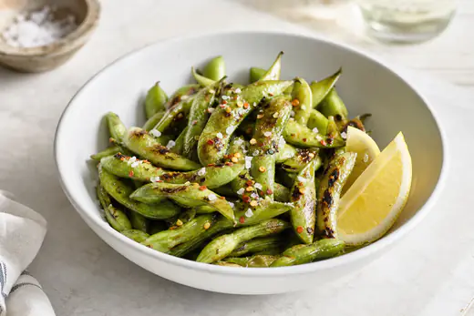 Blistered edamame pods sprinkled with flaky salt and chili flakes in a white bowl