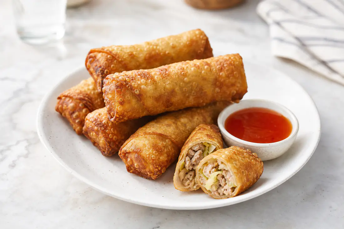 Golden crispy egg rolls, one cut in half to show the pork and cabbage filling, with sweet and sour sauce