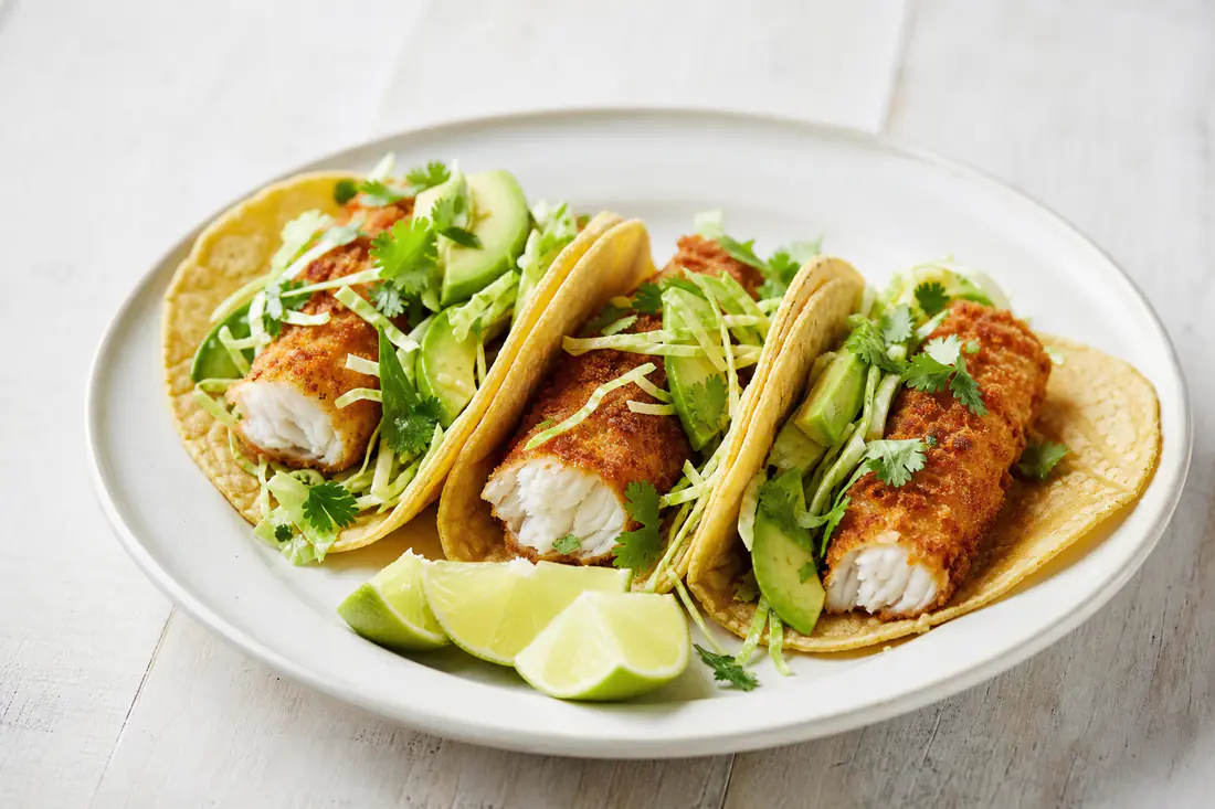 Three crispy fish tacos with cabbage slaw, avocado and lime wedges on a plate