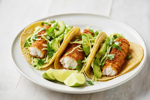 Three crispy fish tacos with cabbage slaw, avocado and lime wedges on a plate