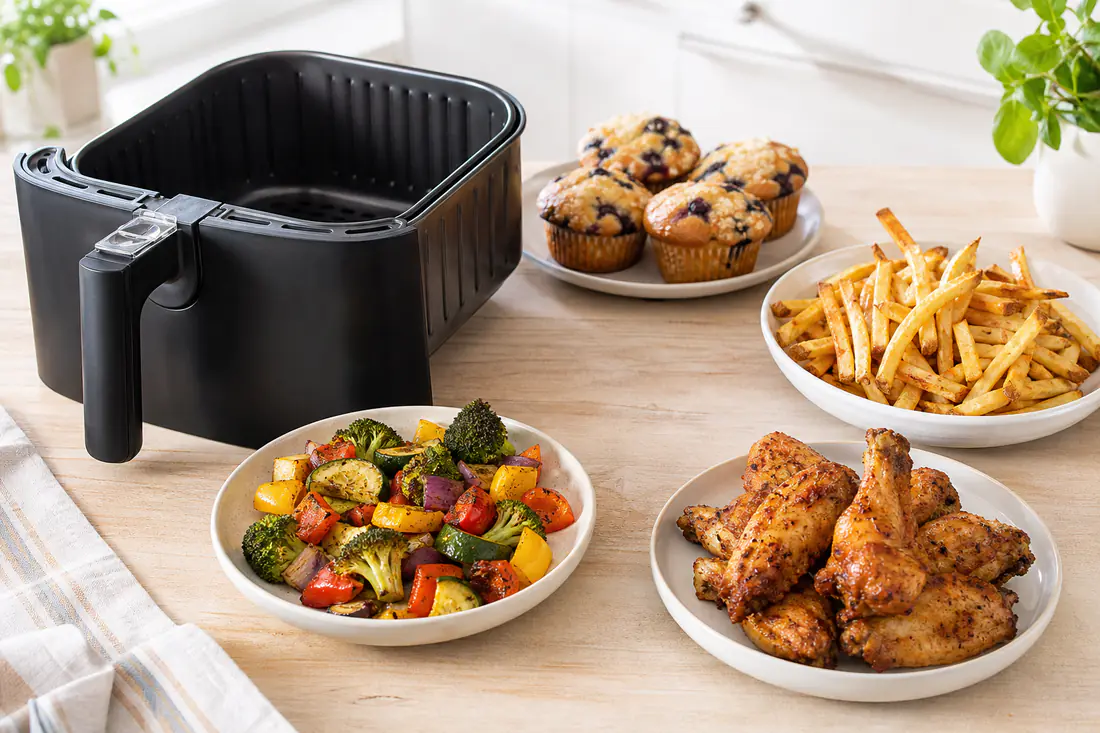 Air fryer on a kitchen counter with french fries, roasted vegetables, chicken wings and muffins