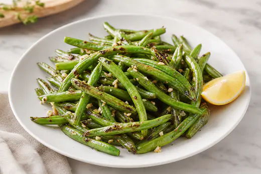 Blistered green beans with garlic and a lemon wedge on a white plate