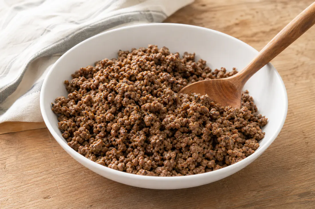 Browned seasoned ground beef crumbles in a white bowl with a wooden spoon