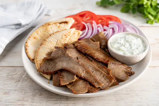 Thin crisp slices of gyro meat on a plate with warm pita bread, tomatoes and tzatziki