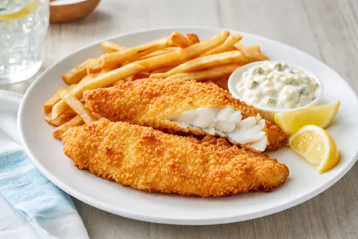 Golden breaded haddock fillets on a plate with lemon wedges, tartar sauce and fries