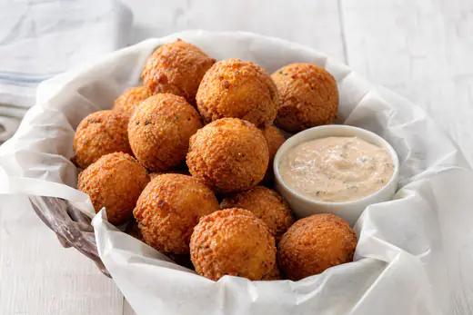 Golden round hush puppies in a paper-lined basket with a small bowl of remoulade sauce
