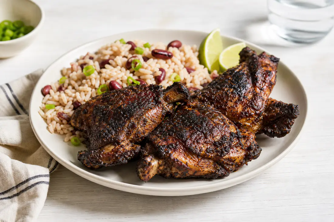 Dark golden jerk chicken thighs with charred spots on a plate with rice and peas and lime wedges