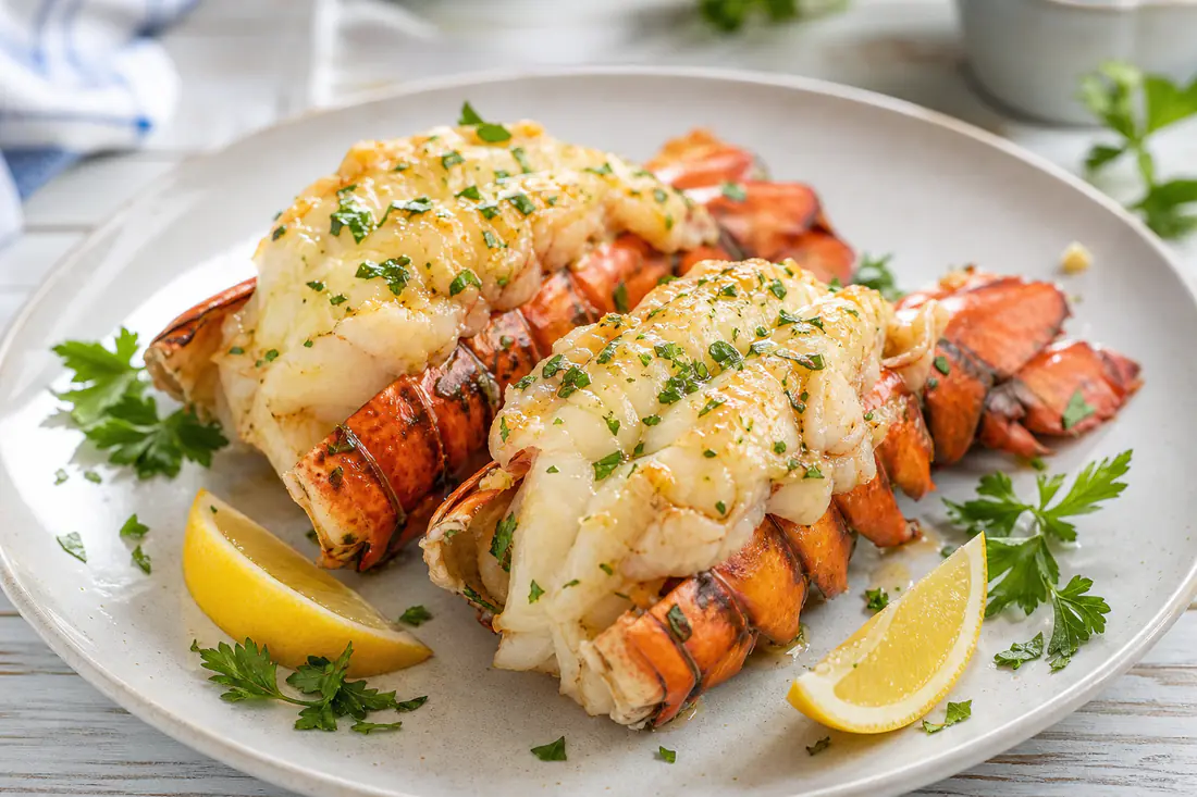 Two butterflied lobster tails in the shell with garlic butter, parsley and lemon wedges