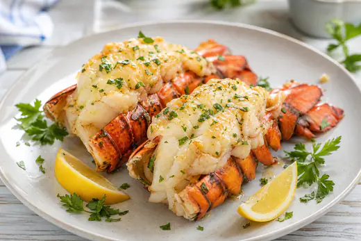 Two butterflied lobster tails in the shell with garlic butter, parsley and lemon wedges