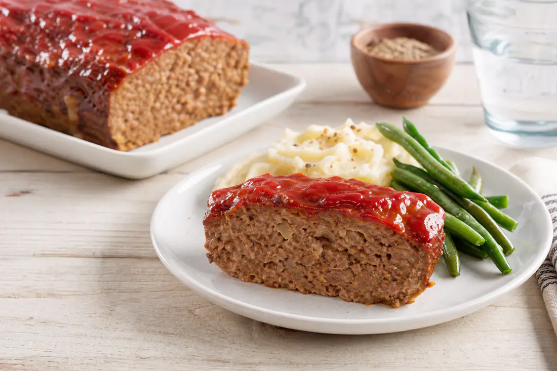 Sliced glazed beef meatloaf with a shiny ketchup topping on a plate with mashed potatoes