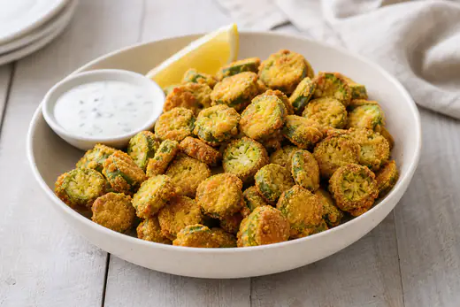 Golden cornmeal-breaded fried okra rounds in a bowl with a small dish of ranch dressing
