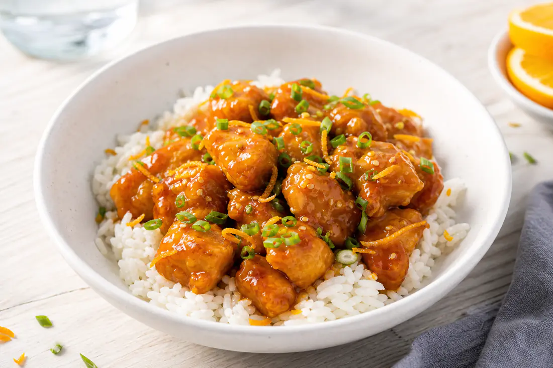 Crispy orange chicken pieces coated in glossy sauce over rice with scallions