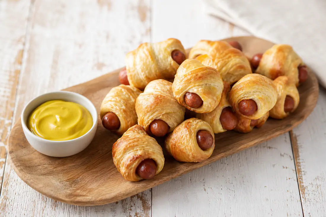 Golden brown mini pigs in a blanket on a serving board with a bowl of mustard