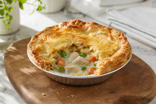 Golden baked chicken pot pie in its small foil pan, with a slice of crust broken to show the filling