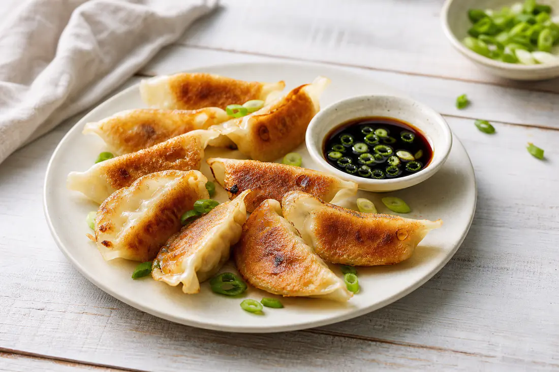 Crisp golden potstickers on a plate with a small bowl of soy dipping sauce and sliced scallions