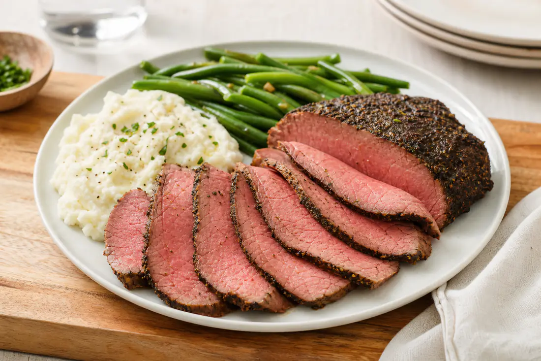 Thinly sliced eye of round roast beef with a seasoned crust and a pink center on a cutting board