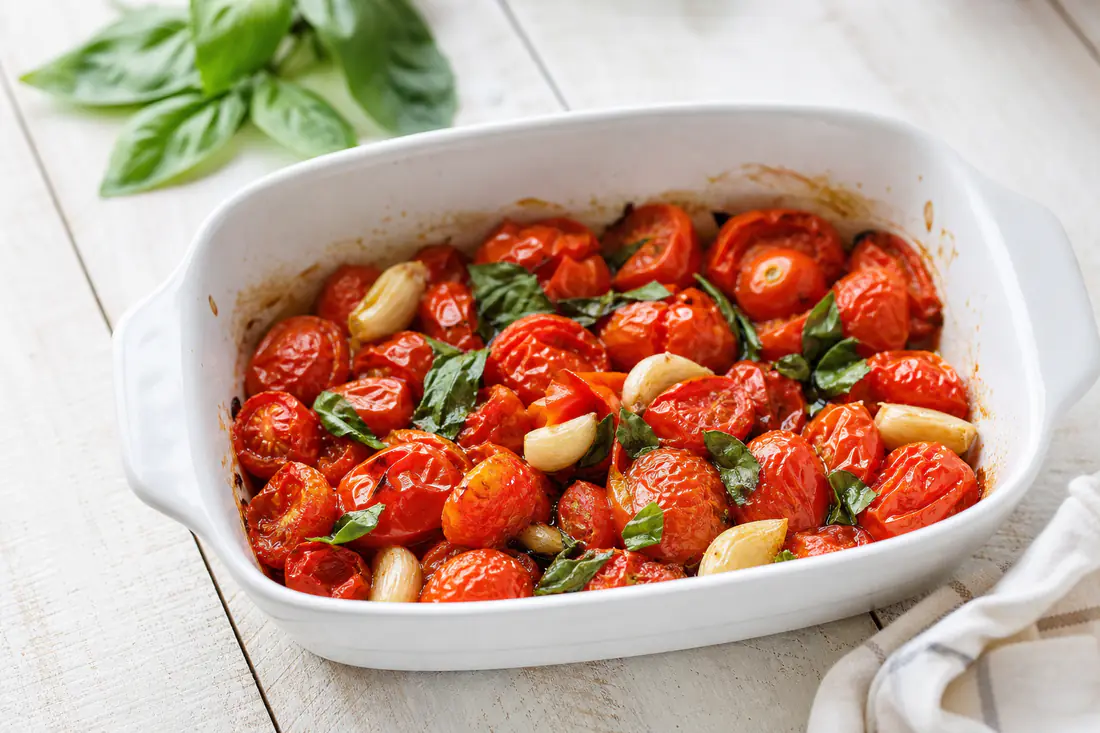 Burst roasted cherry tomatoes with garlic cloves and fresh basil in a white dish