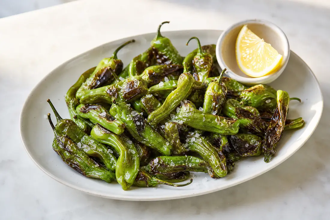 Blistered shishito peppers with flaky salt and a lemon wedge in a small bowl
