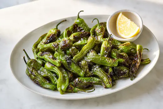 Blistered shishito peppers with flaky salt and a lemon wedge in a small bowl