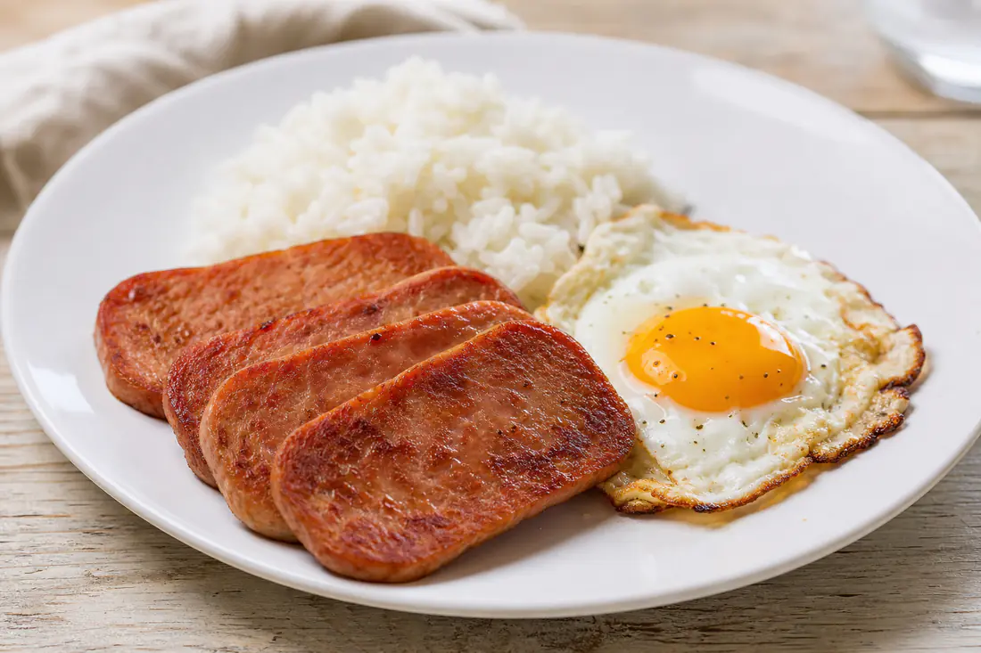 Crispy browned slices of Spam on a white plate beside a fried egg and steamed rice