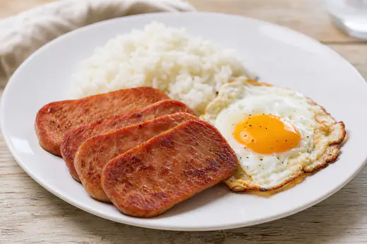 Crispy browned slices of Spam on a white plate beside a fried egg and steamed rice